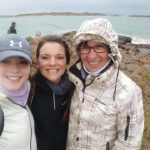 3 women smile for the camera with the coast in the background on a rainy day